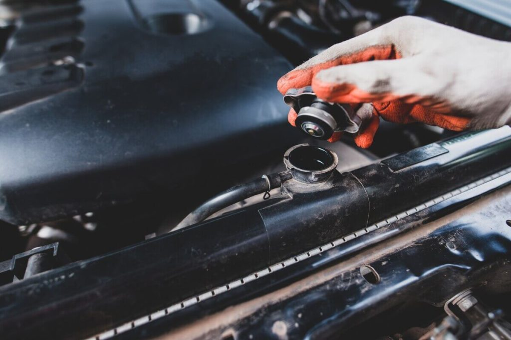 A gloved hand removes a coolant reservoir cap on a car engine.