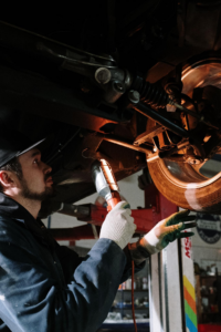 Automotive technician inspecting suspension and wheel components under a raised vehicle with a work light